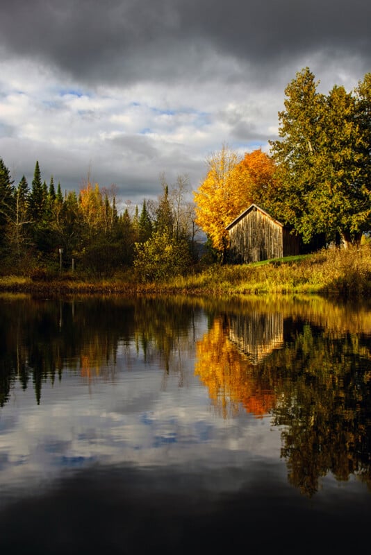 A rustic wooden barn sits beside a calm pond, surrounded by autumn trees under a cloudy, dramatic sky.