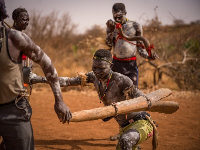 Three men with body paint and traditional adornments participate in a ceremonial activity in a dry, outdoor setting.