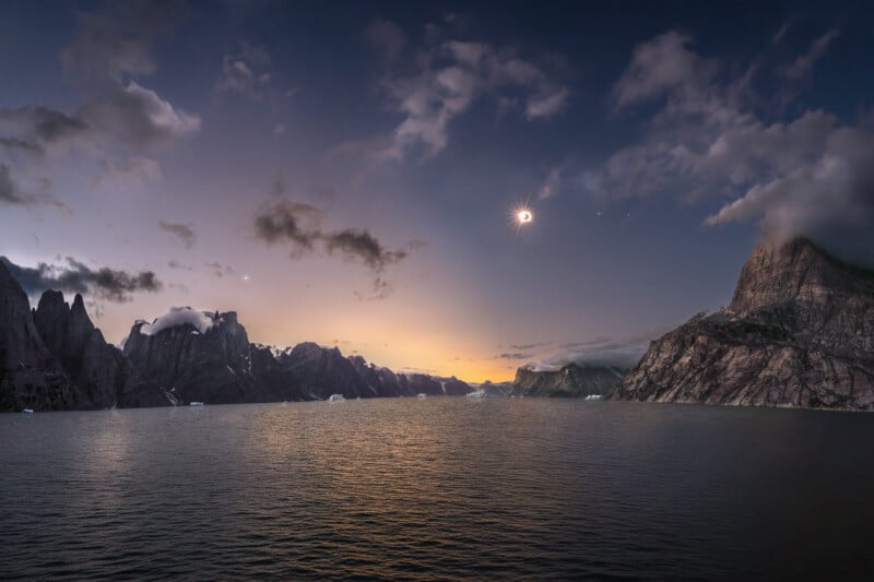 A total solar eclipse hangs in the sky above a calm body of water surrounded by jagged, cloud-capped mountains.