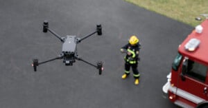 A drone hovers in the air while a firefighter in uniform operates it with a remote control near a red fire truck.