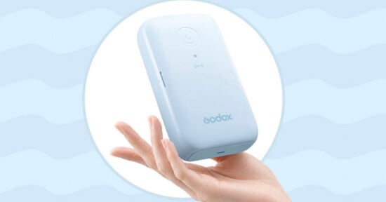 A hand holds a light blue, rectangular Godox device against a light blue background with a wavy pattern.