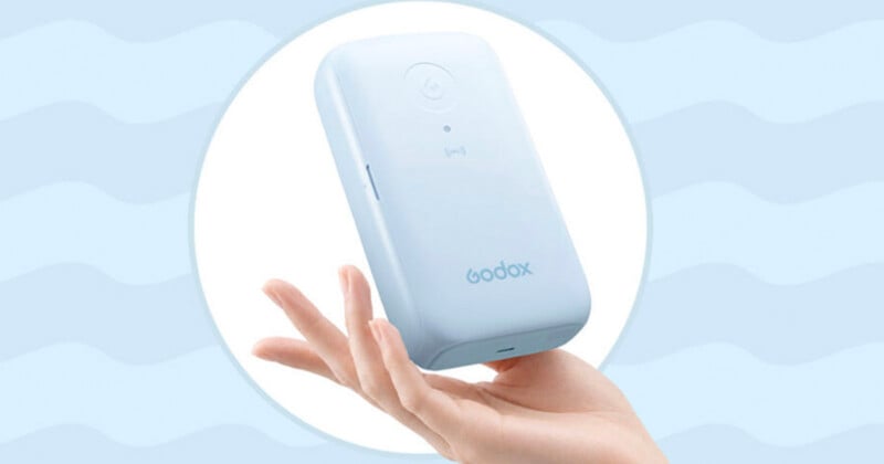 A hand holds a light blue, rectangular Godox device against a light blue background with a wavy pattern.