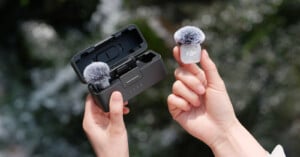 A person holds a small, white wireless microphone with a fuzzy windscreen in their right hand, while their left hand holds an open charging case containing another identical microphone.