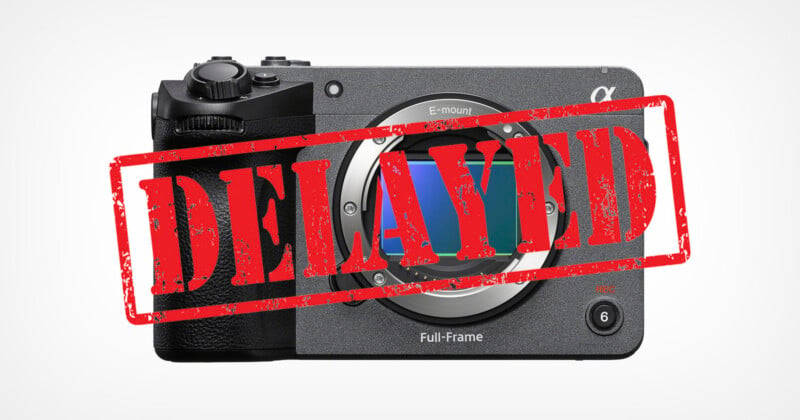 A front-facing view of a gray digital camera body with a lens mount and a red, distressed "DELAYED" stamp overlaid across the center.