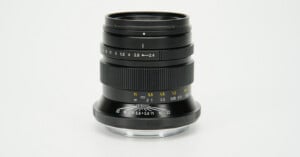 A black manual focus camera lens stands upright against a plain white background, showing aperture and distance markings.