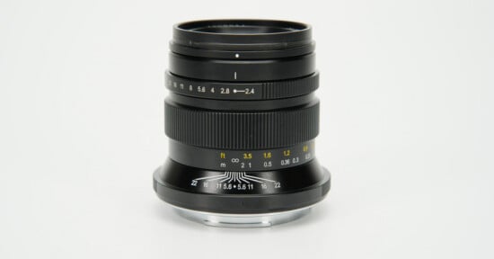 A black manual focus camera lens stands upright against a plain white background, showing aperture and distance markings.
