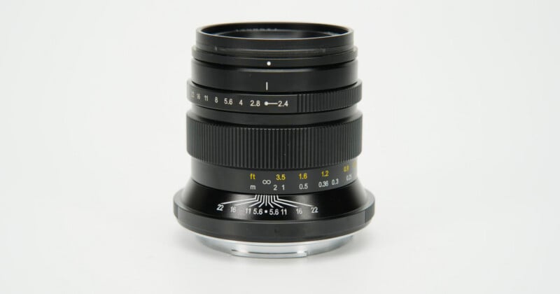 A black manual focus camera lens stands upright against a plain white background, showing aperture and distance markings.