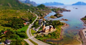 A large, light-colored church with a tall spire sits on a coastal point next to a road, small houses, and a calm, blue bay.