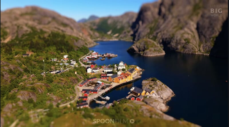 A tilt-shift aerial view of a small coastal village with colorful houses nestled between rocky mountains and a deep blue fjord.