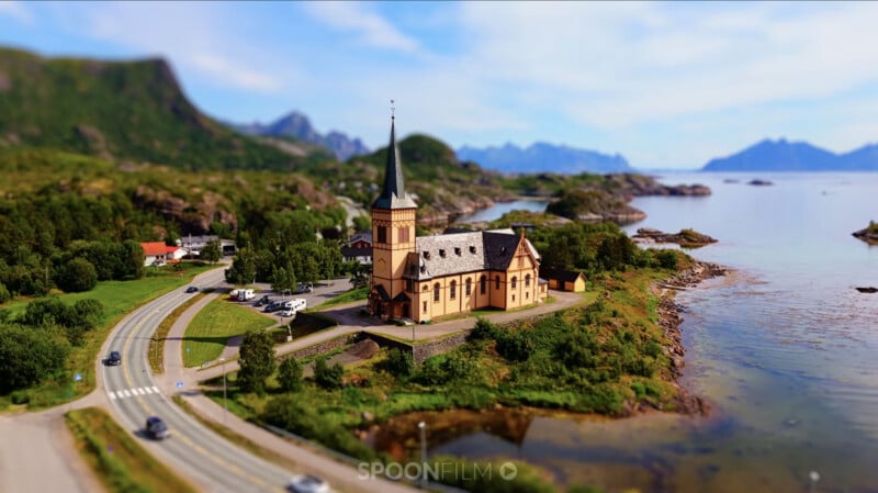 A yellow church with a tall steeple stands on a grassy coastal point next to a winding road and a calm body of water.