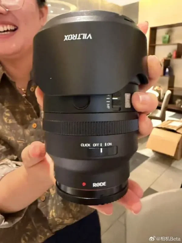 A person holds a black camera lens with a Viltrox lens hood and a Rode label near the mount.