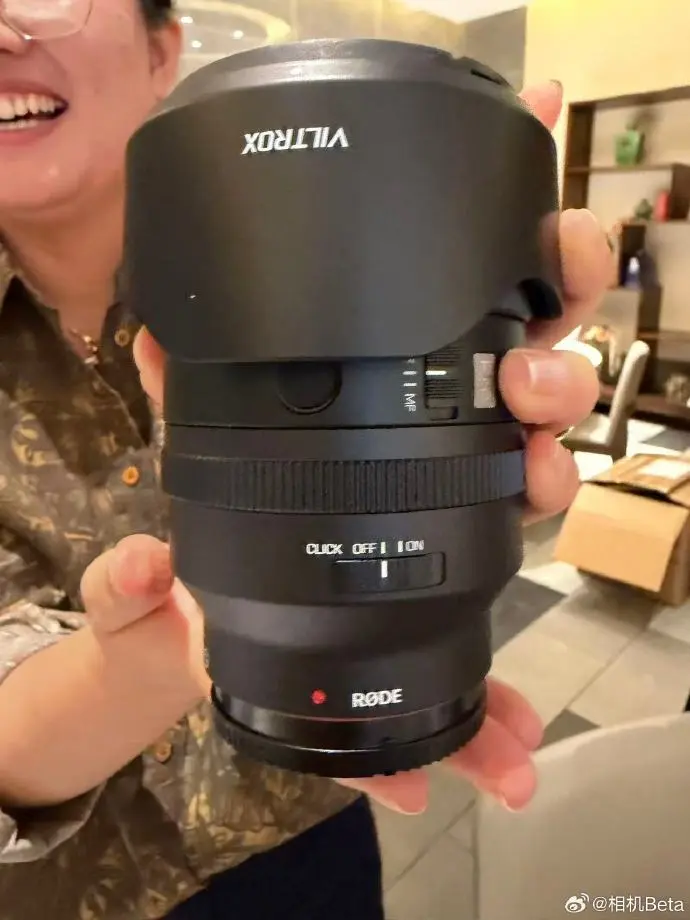 A person holds a black camera lens with a Viltrox lens hood and a Rode label near the mount.