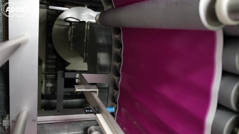 A magenta-colored film strip moves vertically through a series of industrial rollers in a manufacturing machine.