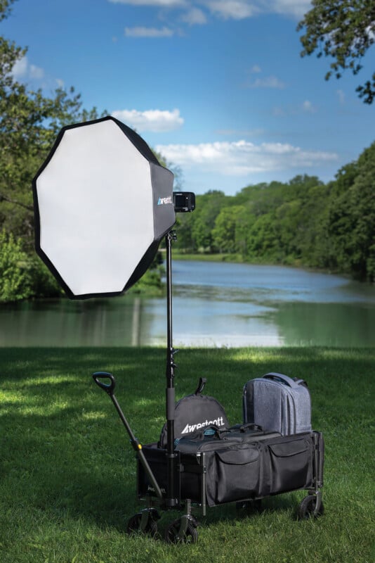 A Westcott octagonal softbox on a light stand is mounted inside a collapsible utility wagon containing camera bags on a grassy lawn near a river.