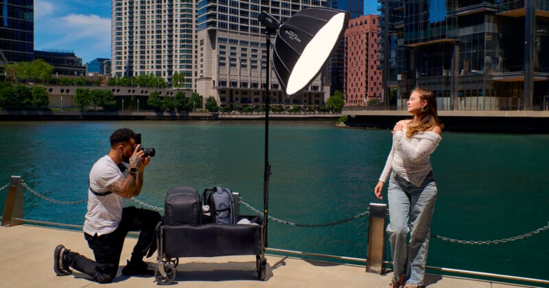 A photographer kneels on a concrete walkway to take a picture of a woman posing near a river, with a large portable light stand and equipment cart positioned between them.