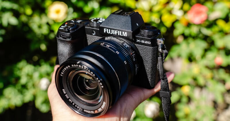 A hand holds a black Fujifilm X-S20 mirrorless camera with an 18-55mm lens attached, set against a blurred background of green foliage and flowers.