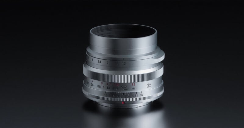 A silver manual focus camera lens with aperture and distance markings sits against a dark, reflective background.