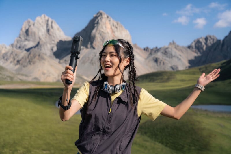 A smiling woman with braided hair and sunglasses on her head holds a 360-degree camera on a selfie stick while standing in a mountain landscape.