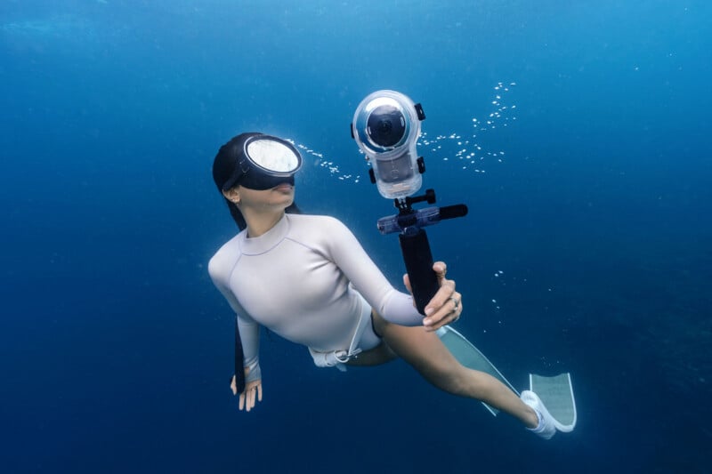A person in a white swimsuit and diving mask swims underwater while holding a 360-degree camera on a handle.