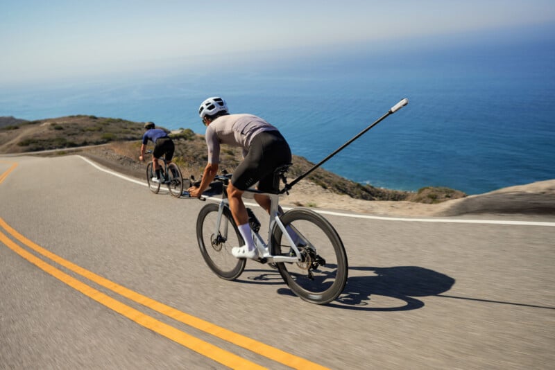 Two cyclists ride road bikes along a winding coastal highway with the ocean in the background, one with a camera mounted on an extended pole.
