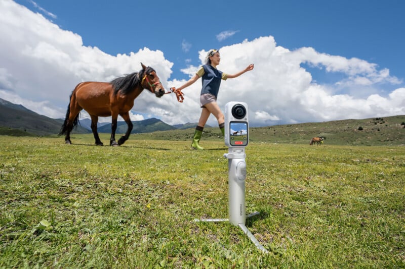 A white 360-degree camera on a tripod stand sits in a grassy field, recording a woman walking a brown horse in the background.
