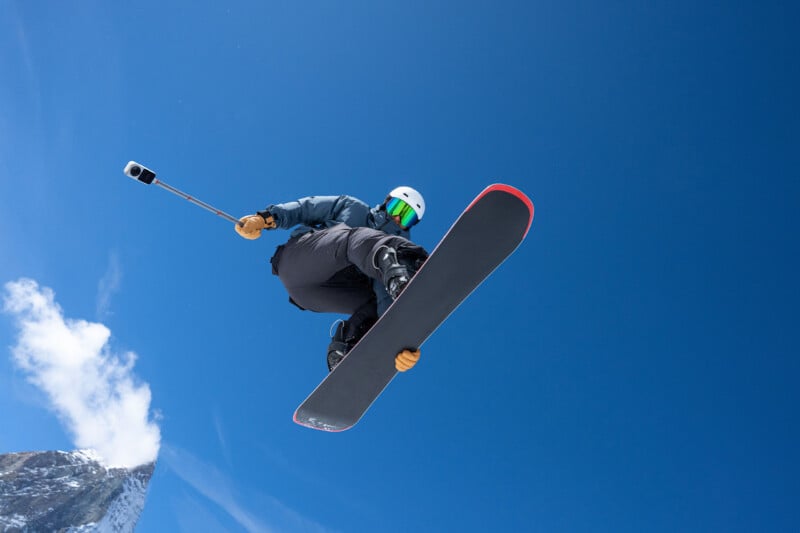 A snowboarder in a blue jacket and white helmet jumps against a clear blue sky while holding a camera on an extension pole.