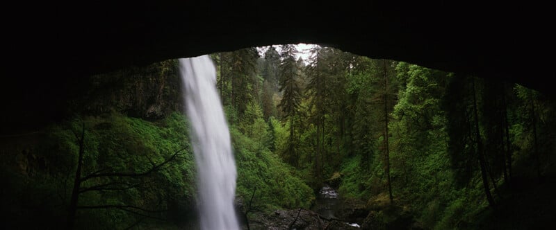 A waterfall cascades down a lush, green forested canyon, viewed from beneath a dark rock overhang.