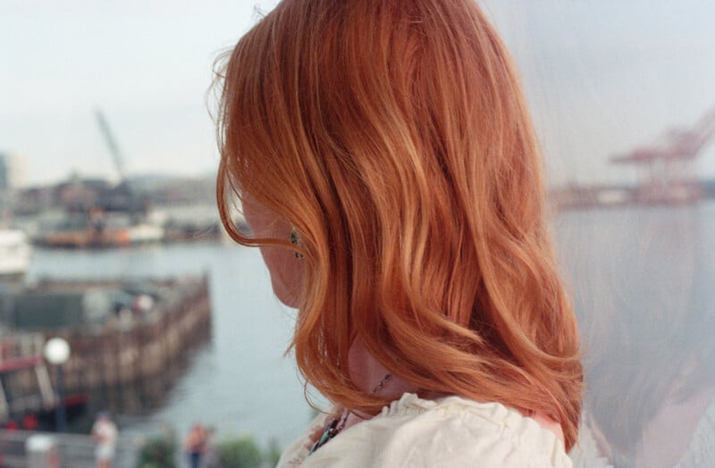 A person with shoulder-length wavy red hair is seen in profile, looking out toward a harbor with industrial cranes.