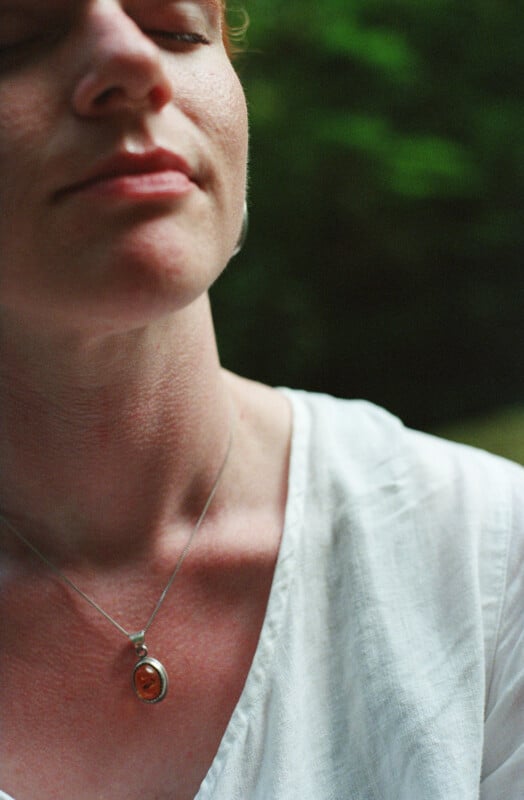 A close-up shot of a person with fair skin and red hair, eyes closed, wearing a white linen shirt and an amber pendant necklace.