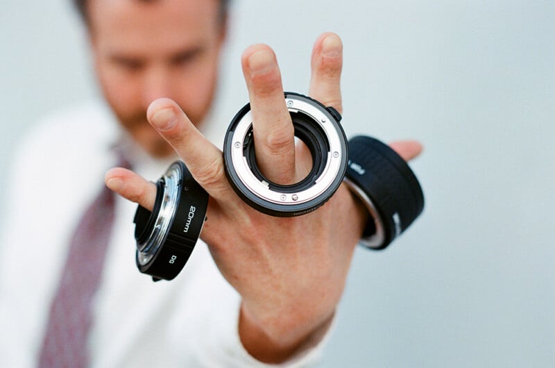 A person wearing a white shirt and tie holds three camera extension tubes on their fingers, with one labeled "20mm" visible.