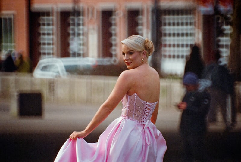 A blonde woman in a strapless pink gown with a corset back looks over her shoulder while holding the edge of her skirt.