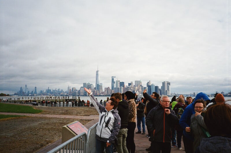 A group of people stands along a waterfront railing, with one woman pointing toward the distant city skyline under a cloudy sky.