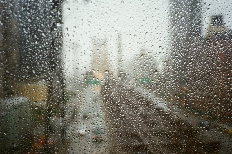 Raindrops cover a window, blurring the view of a city street with buildings and traffic below.