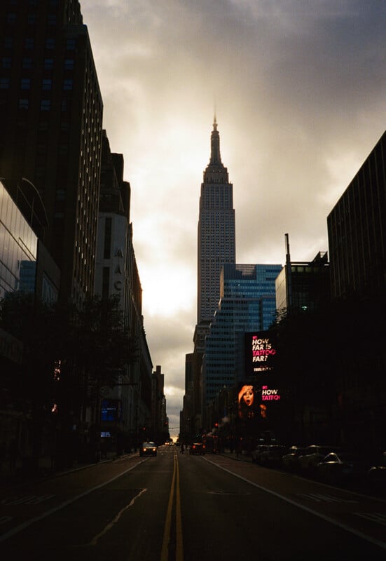 A street-level view looking down a city avenue toward the Empire State Building under a cloudy, backlit sky.