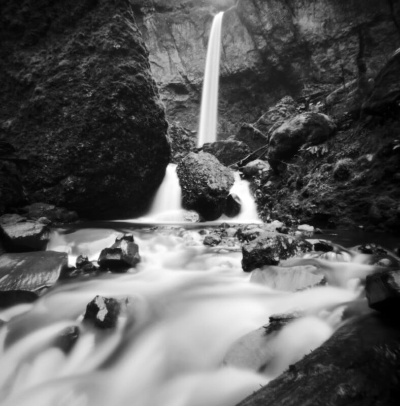 A black and white long-exposure shot of a waterfall cascading over rocky cliffs into a flowing stream.