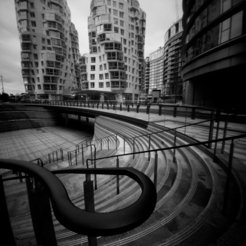 A black and white view of curved concrete stairs leading down to a plaza, framed by modern high-rise apartment buildings.
