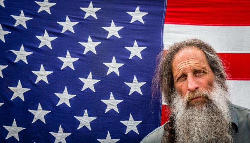 A man with long gray hair and a full beard stands in front of a large American flag.