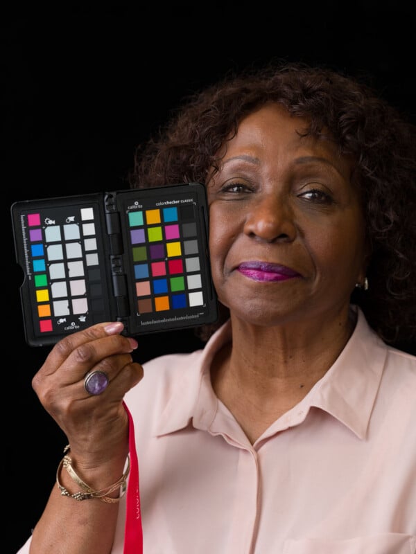 A woman with curly dark hair and a light pink shirt holds a Calibrite ColorChecker Classic card next to her face.
