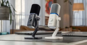 Two amaran microphones, one black and one white, stand on a desk next to laptops in a brightly lit room.