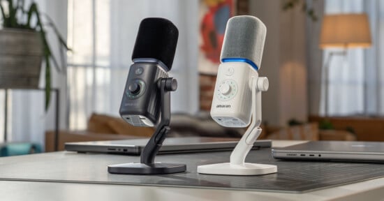 Two amaran microphones, one black and one white, stand on a desk next to laptops in a brightly lit room.