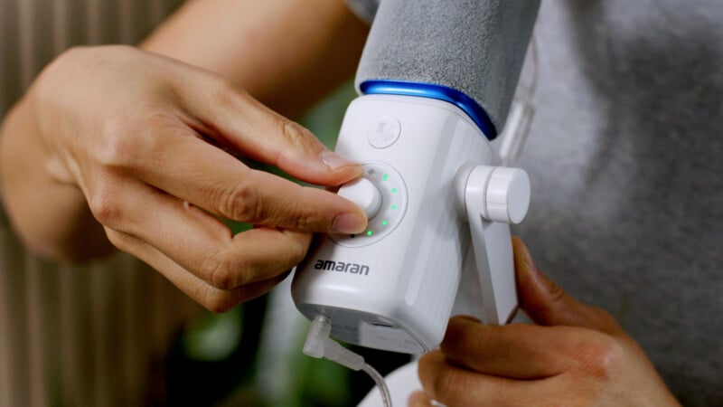A person's hands adjust the dial on a white Amaran brand lighting device, which features a circular LED indicator and a power cable connected to the base.