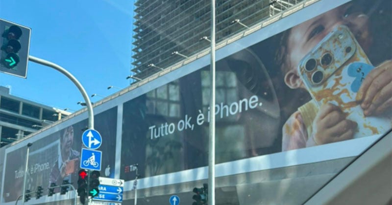 A large billboard displays an advertisement featuring a child holding a messy iPhone with the text "Tutto ok, è iPhone." above a busy city intersection with traffic lights.