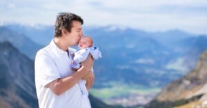 A man in a white polo shirt kisses the forehead of a newborn baby he is holding against a backdrop of mountain ranges.