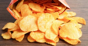 A bag of ridged potato chips lies on its side on a wooden surface, with many chips spilled out in front of it.