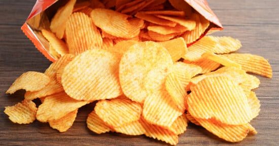 A bag of ridged potato chips lies on its side on a wooden surface, with many chips spilled out in front of it.