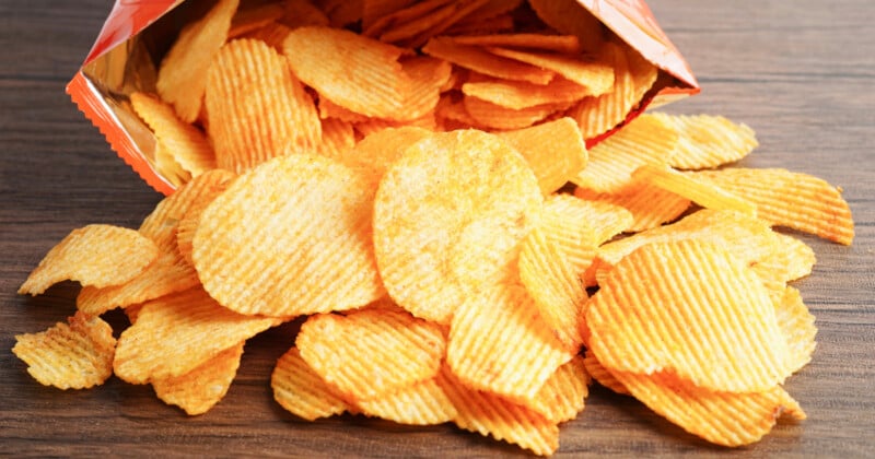 A bag of ridged potato chips lies on its side on a wooden surface, with many chips spilled out in front of it.