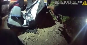 A body-worn camera view shows a person reaching into an open black backpack resting on a rocky, dark ground at night.