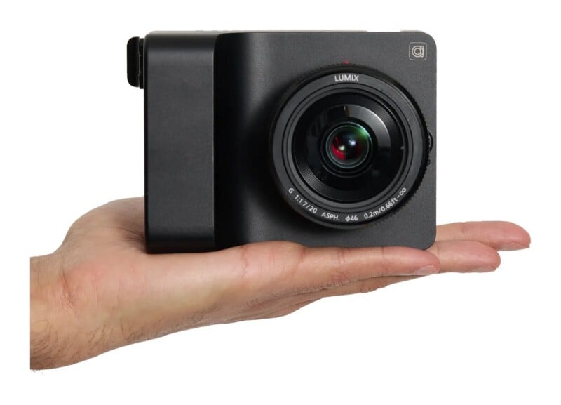 A hand holds a small, black, box-shaped camera with a prominent Lumix lens attached to the front.
