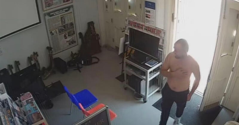 A shirtless man stands in the doorway of a room containing a television on a metal cart, two chairs, and various posters and objects on the walls.