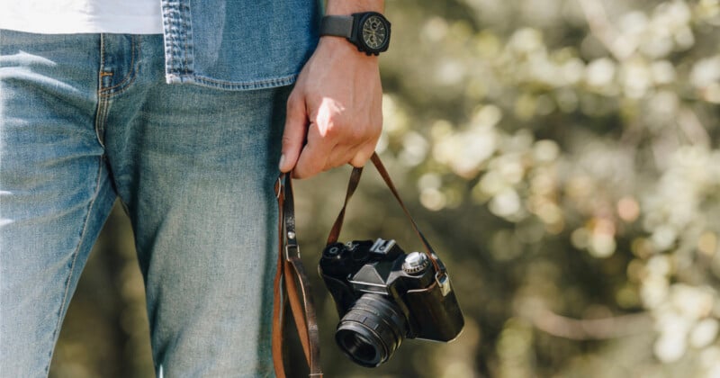 A person wearing a denim shirt and jeans holds a vintage film camera by its leather strap, with a watch visible on their wrist.